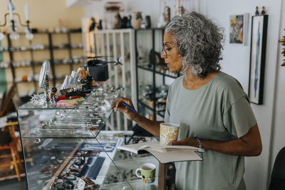 Senior female entrepreneur with diary and coffee cup examining jewelry at antique shop
