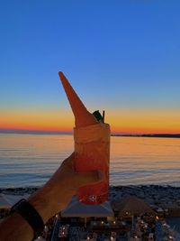 Hand holding drink at beach against sky during sunset