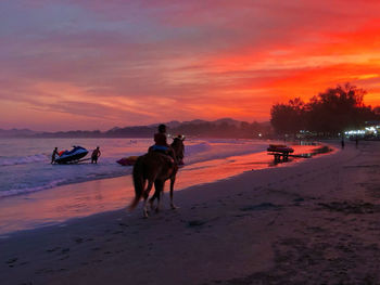 People riding horse on beach against sky during sunset