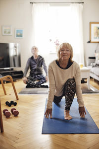 Smiling senior woman practicing yoga on exercise mat with friend in background at home