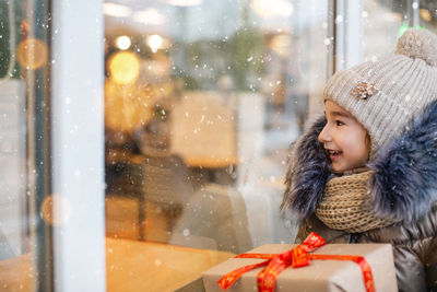 Cute smiling girl with christmas gift looking through window
