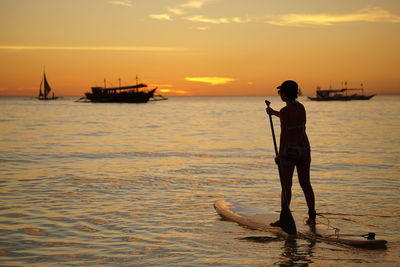 Silhouette woman paddleboarding in sea against orange sky