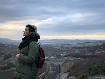 Side view of young woman with backpack standing on mountain against cloudy sky during sunset