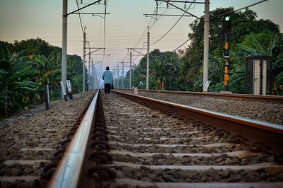 Rear view of man walking on railroad tracks against sky during sunset