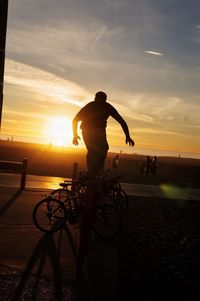 Silhouette man riding bicycle by sea against sunset sky