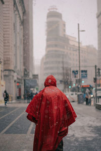 Back view of anonymous male in bright red protective cloak walking on street with buildings in snowy winter in madrid spain