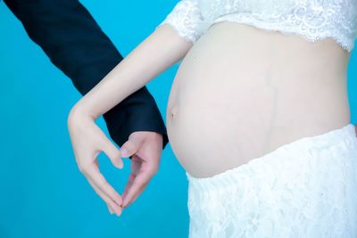 Midsection of pregnant woman making heart shape with man against blue background