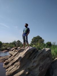 Low angle view of mature man standing on rock against blue sky