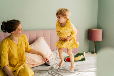 A small child girl jumps cheerfully on the bed, mom laughs and sits next to her.