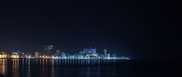 Illuminated buildings by sea against clear sky at night