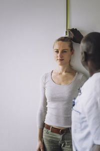 Doctor measuring height of female patient standing against wall in clinic