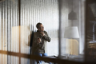 Mature businessman talking on smart phone through headphones seen from glass