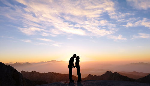 Silhouette man kissing woman while standing against cloudy sky during sunset