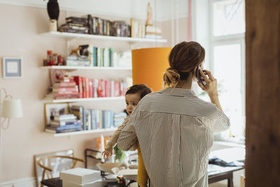 Rear view of businesswoman carrying baby boy while talking on smart phone at home