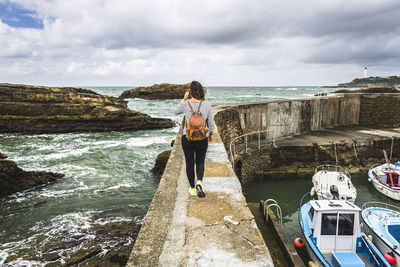 Rear view of young woman standing with backpack walking at beach against cloudy sky