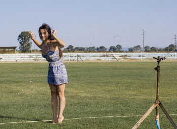Full length of smiling young woman standing on football field, playing with sprinkler