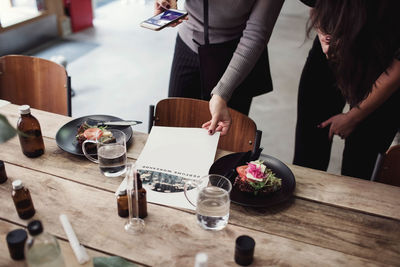 Midsection of multi-ethnic female colleagues photographing brochure with food and perfume on table at workshop