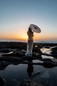 Man standing on rock at beach against sky during sunset