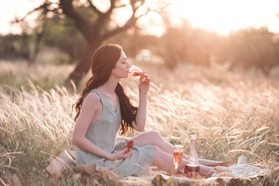 Stylish woman drinking wine with strawberry siting in field with picnic outdoor at sunset. 