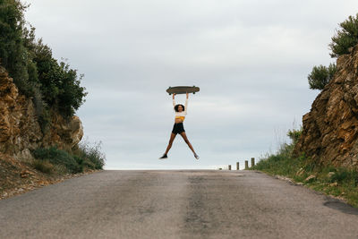 Full body of positive ethnic female jumping high and raising hands with longboard on asphalt road