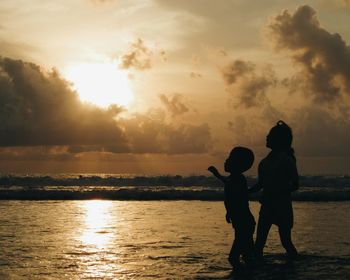 Silhouette mother and daughter standing by sea against sky during sunset