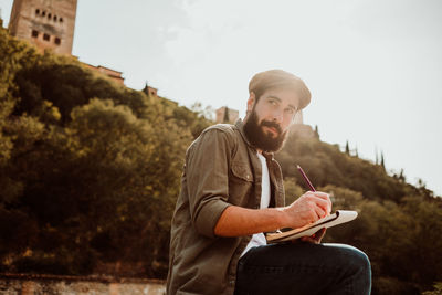 Thoughtful man holding book and pencil while sitting against sky