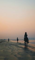 People riding horses on beach against sky during sunset