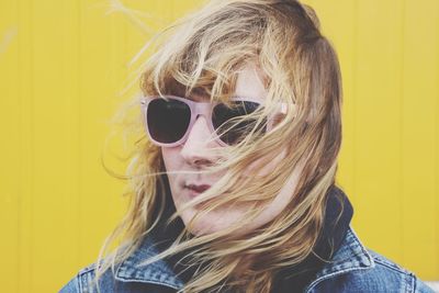 Close-up of woman with tousled hair wearing sunglasses against yellow wall