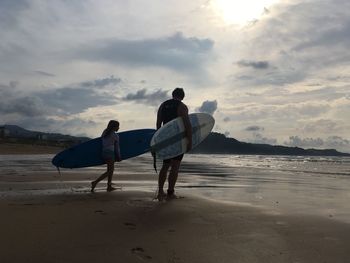 Father and daughter with surfboard walking at beach against sky