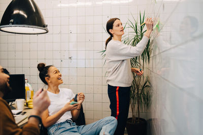 Smiling colleagues looking at young businesswoman writing strategy on tile wall in creative office