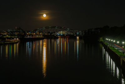 Illuminated city by river against sky at moonrise at night
