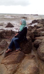 Woman sitting on rock at beach against sky