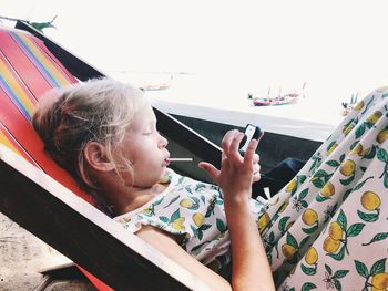 Girl with lollipop using phone while sitting on deck chair