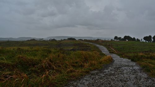 Dirt road on field against cloudy sky