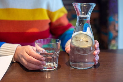 Women's hands hold a pitcher of water and lemon next to a glass glass . multicolored sweater