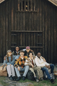 Portrait of happy multiracial male and female friends sitting with arm around each other in front of log cabin