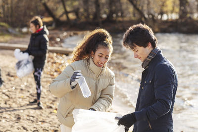 Smiling female volunteer with male friend holding plastic bottle in garbage bag