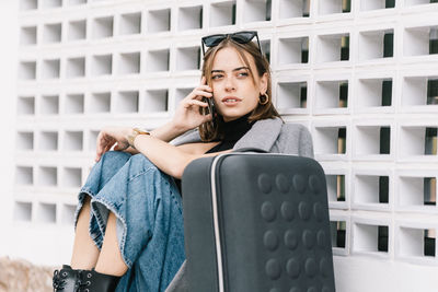 Female tourist with luggage sitting alone on pavement and speaking on mobile phone while getting lost in city looking away