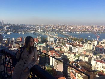 Woman leaning on railing of galata tower by golden horn metro bridge and cityscape against clear blue sky