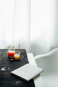 From above of jar and glass with red and orange fruit drinks placed with book on table near window in light room