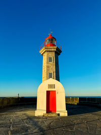 Lighthouse by sea against clear blue sky