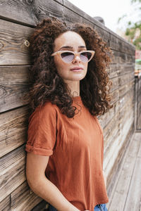 Beautiful young woman with curly hair wearing sunglasses leaning wooden wall