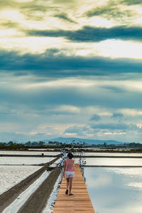 Rear view of mid adult woman walking on pier over lake against cloudy sky during sunset