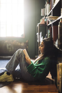 Side view of girl reading book while reclining on shelf in bookstore