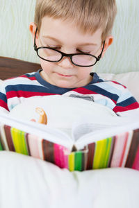 Cute boy reading book while sitting on bed at home