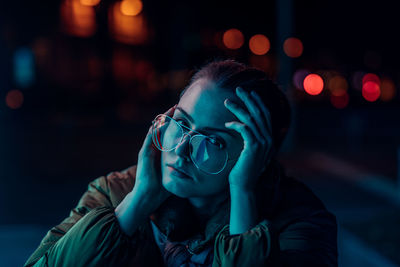 Portrait of young woman wearing eyeglasses against illuminated lights at night