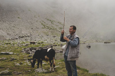 Man holding stick with cow grazing on land by lake against mountain