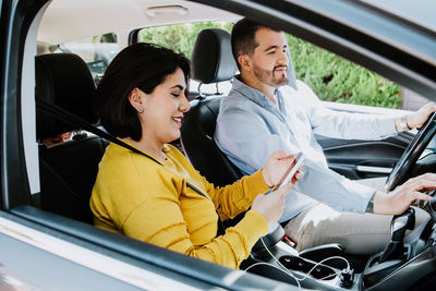 Side view of cheerful young latin american woman with dark hair in casual clothes laughing while using smartphone sitting in car with happy husband