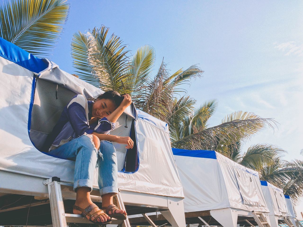 low angle view, tree, palm tree, waist up, sky, clear sky, person, casual clothing, young adult, looking at camera, cloud - sky, day, blue, focus on foreground, exploration