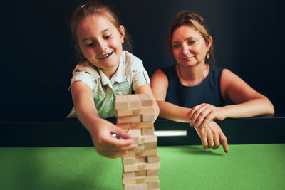 Mother and her daughter playing jenga game together in play room. girl removing one block from stack